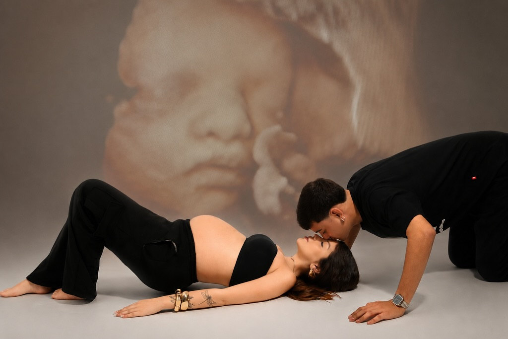 Casal deitado no chão do estúdio se beijando, com a imagem do ultrassom do bebê projetada ao fundo