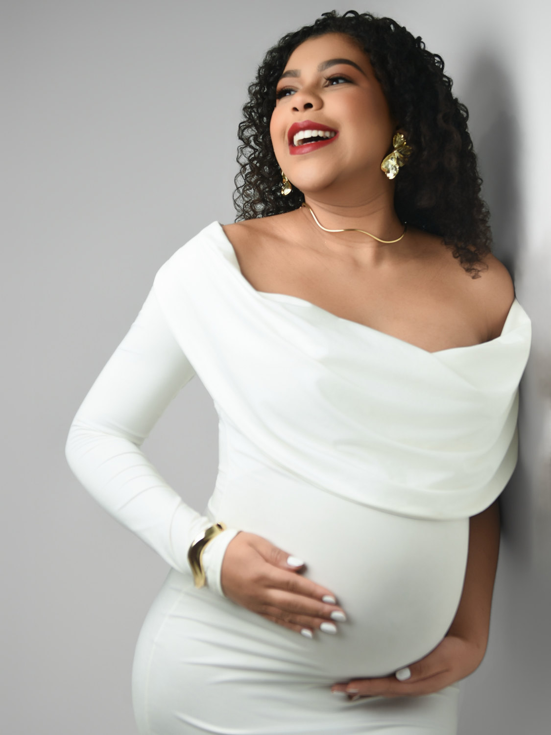 Gestante de vestido branco sorrindo, encostada na parede